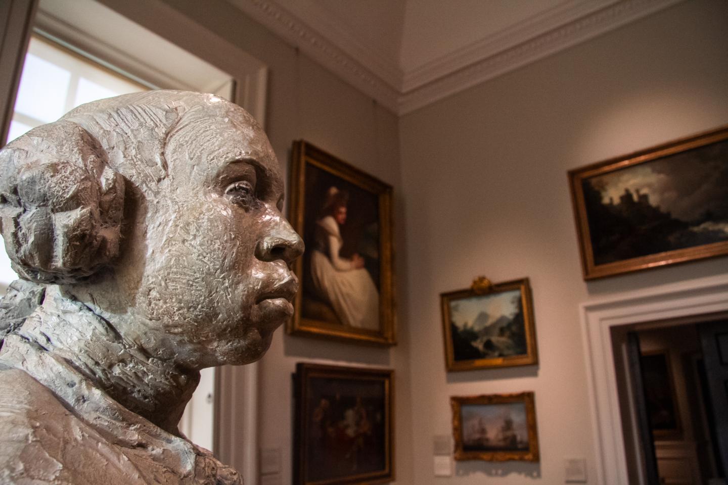 Sculpture of a man viewed from the side, with paintings on display on the wall in the background
