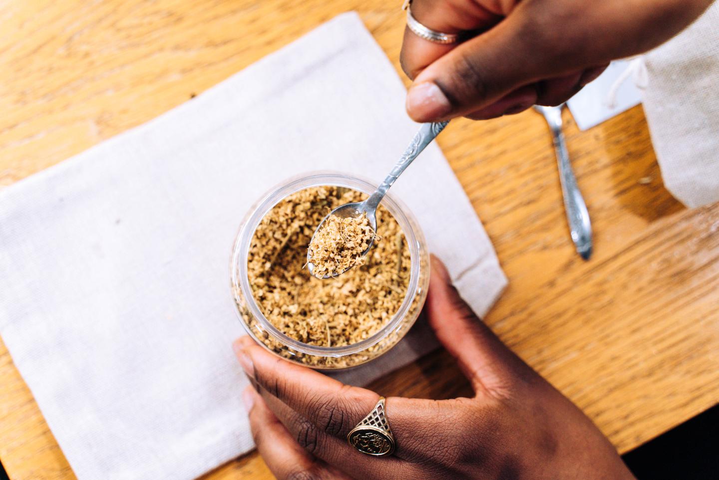 A picture of a hand holding a pot of herbal tea, whilst the other hand spoons out the tea