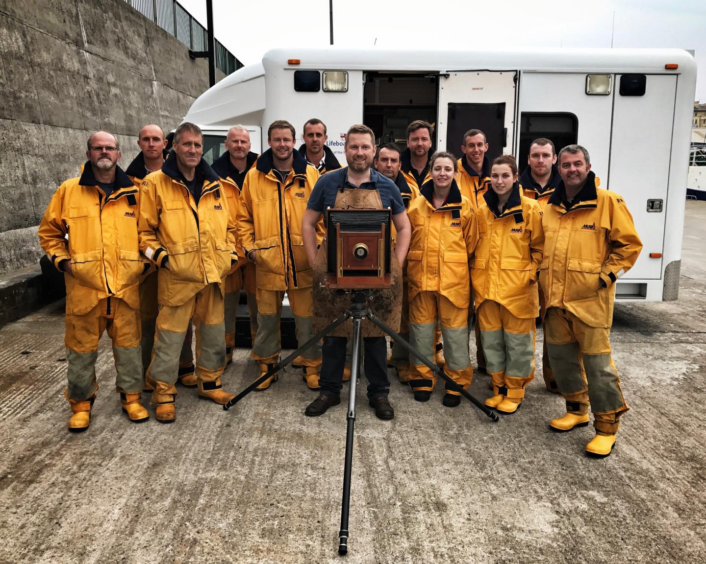 A man with an Edwardian camera stands in the middle of RNLI crew wearing yellow jackets