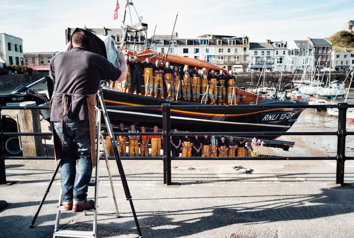 A man stands beneath a focus cloth of an Edwardian camera taking a picture of crew in front of a lifeboat