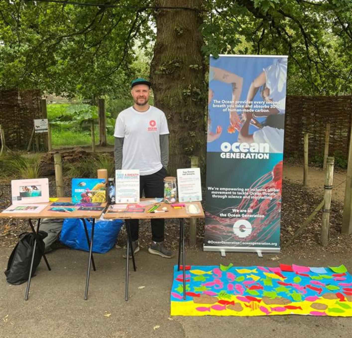 A person stands in front of a stand in a park with an Oceans Generation banner