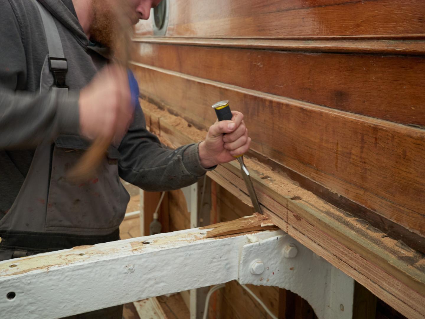 A shipwright is hitting a chisel on a ship plank