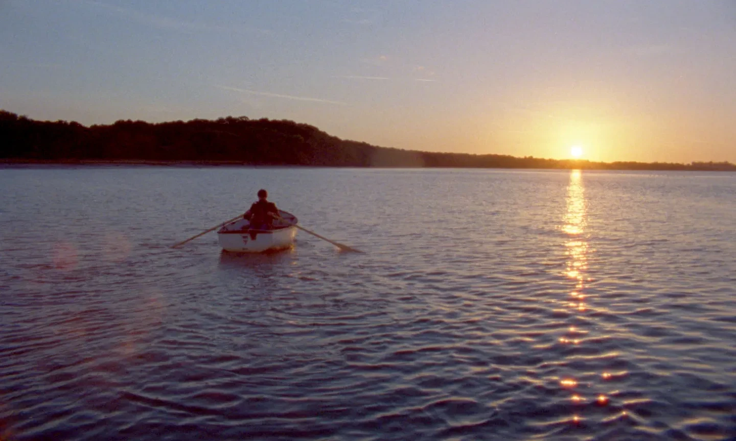 A man in a small rowing boat on calm water, with trees and a sunset in the background.