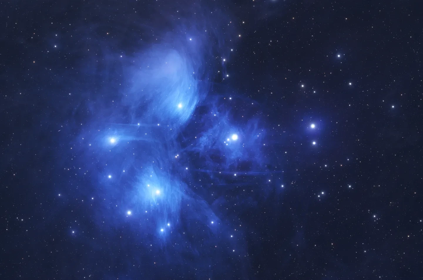 Image of tens of bright light blue stars surrounded by a cloud of gas and dust in varying shades of blue. Behind them is a black starry sky.