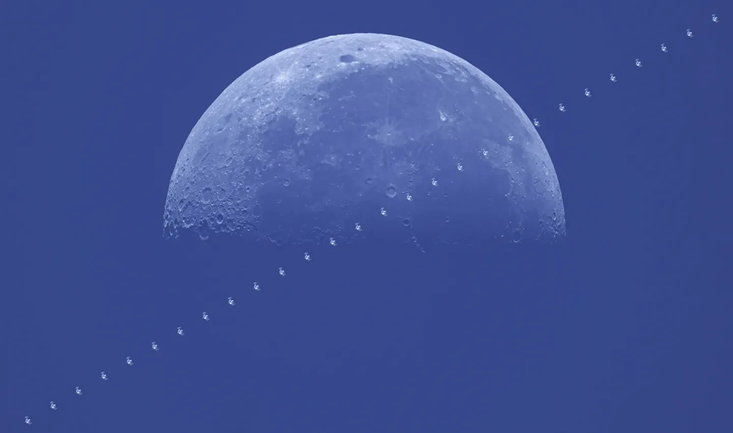 Image of half of a full Moon with a blue sky behind it. Across it diagonally is a series of dark 'H' shapes which shows the passing of the International Space Station. 