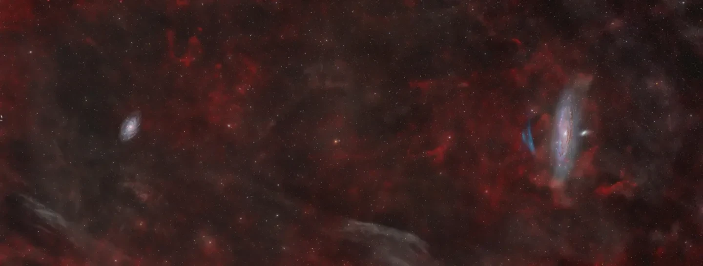 Long letterbox-shaped photograph showing thick clouds of red gas and dust in the sky, covering the whole of the image. On the left side of the image is a small spiral galaxy which has a yellow core and wispy blue and purple spiral arms spiralling around it. On the opposite of the image is the Andromeda Galaxy, side on and vertical in blue and pink spirals around a white core. To its left is a blue oxygen nebula in a 4 shape