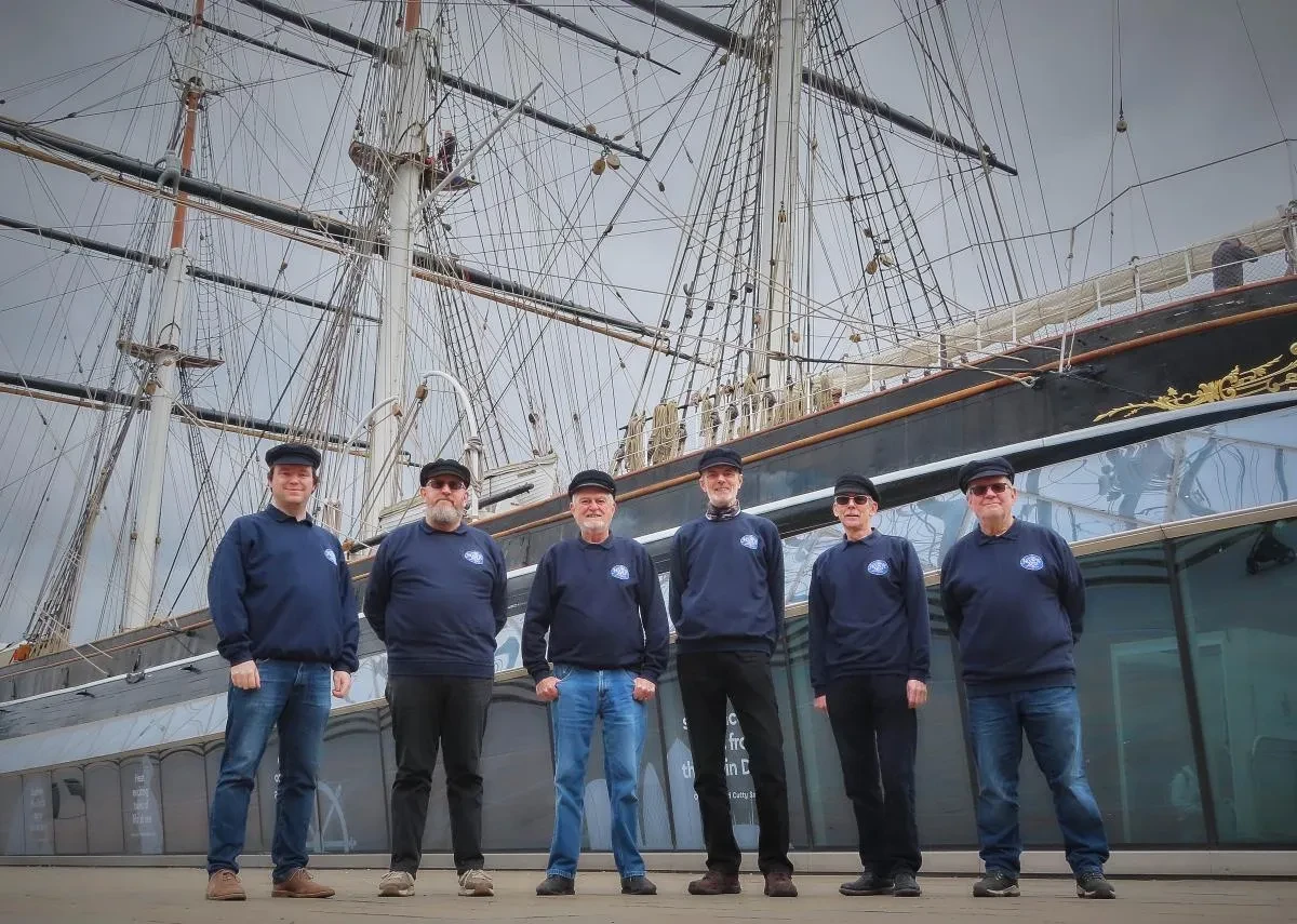 A group of male singers wearing navy jumpers and caps stand in front of Cutty Sark
