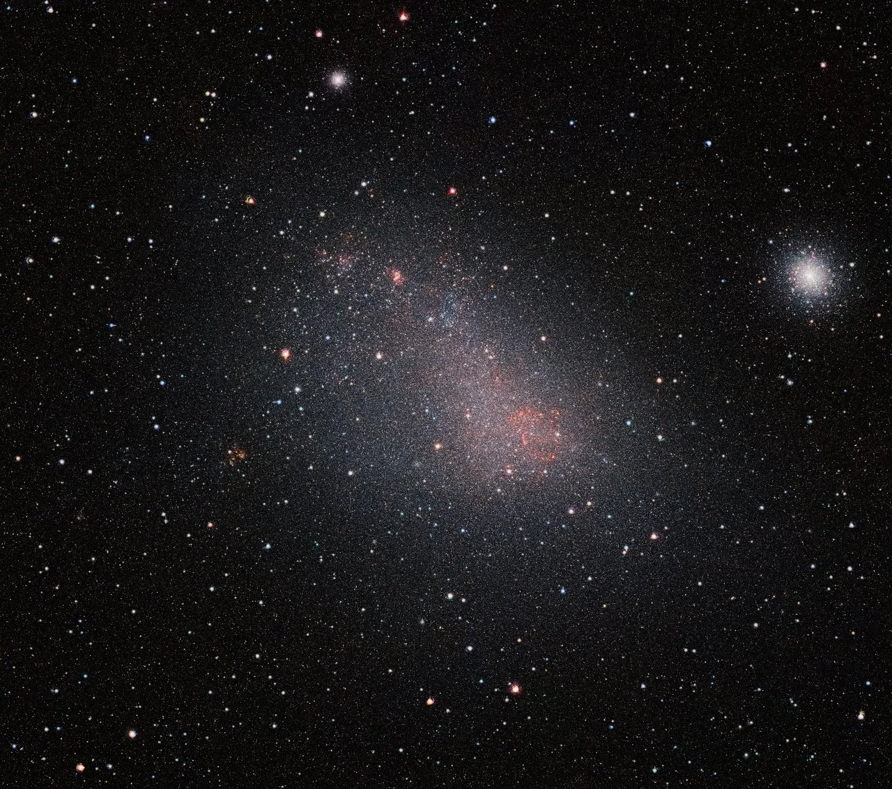 Image of the Small Magellanic Cloud which appears as a haze of white, pink and red light against a black starry sky
