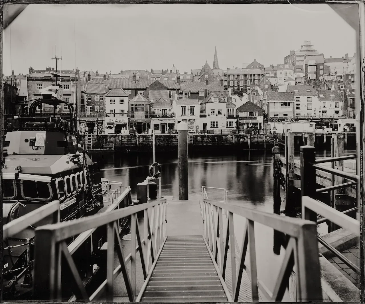 Black and white photography of the view from the gangway at Whitby Lifeboat Station. Buildings line the harbour on the far side, and boats are visible next to the slipway