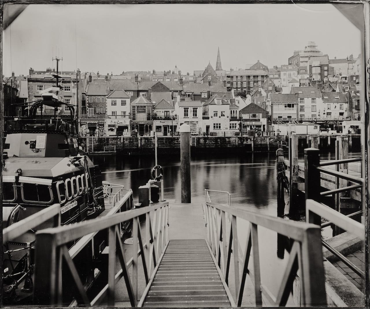 Black and white photography of the view from the gangway at Whitby Lifeboat Station. Buildings line the harbour on the far side, and boats are visible next to the slipway