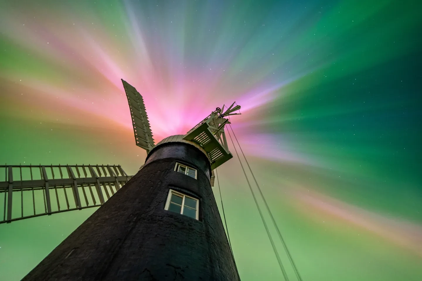 The dark silhouette of a windmill seen from below, with a view of the aurora filling the sky above. The pinkish blue and green light appears to emanate from the top of the windmill