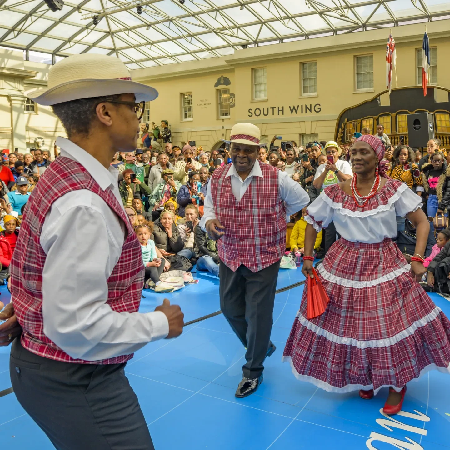 A performance of quadrille dancing at the National Maritime Museum. Two men and a woman are in the centre, dressed in red and white chequered clothes, as a large crowd watches their routine