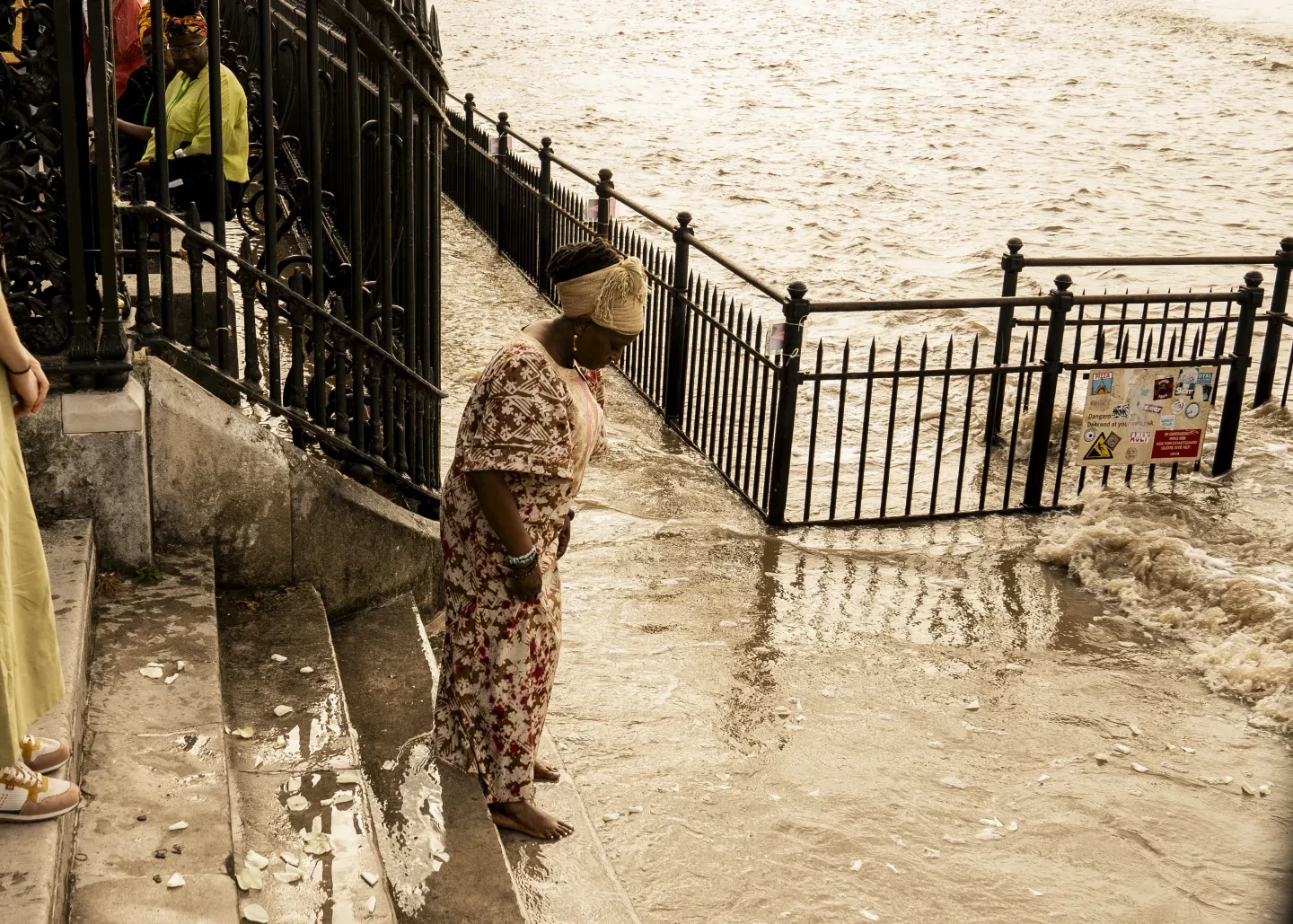 A woman stands by the River Thames at high tide. The river has flooded the Thames footpath, and water is lapping against the stairs the woman is standing on