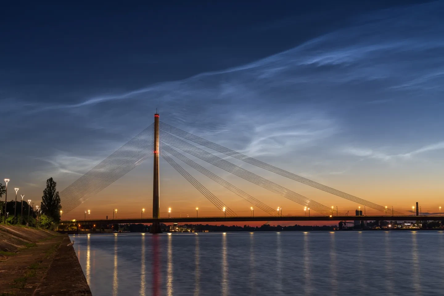 Wispy clouds in the night sky above a suspension bridge on a wide river.
