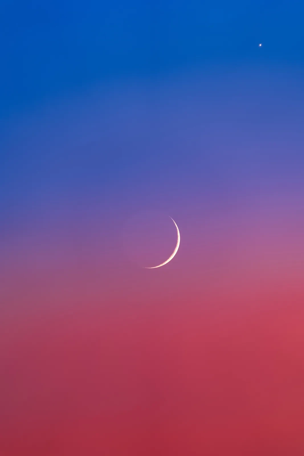 Photo showing thin crescent Moon against a sky that phases from indigo at the top to pink at the bottom