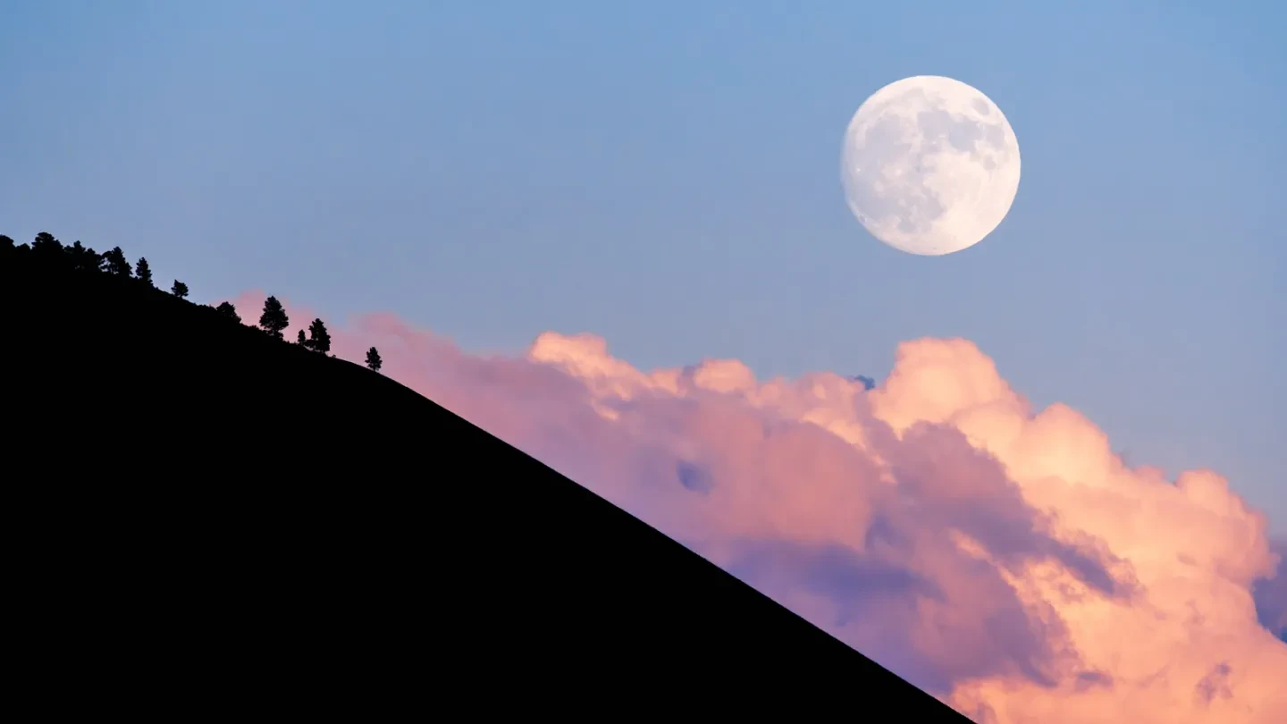 The moon rising in a blue cloudy sky above the silhouetted side of a mountain.