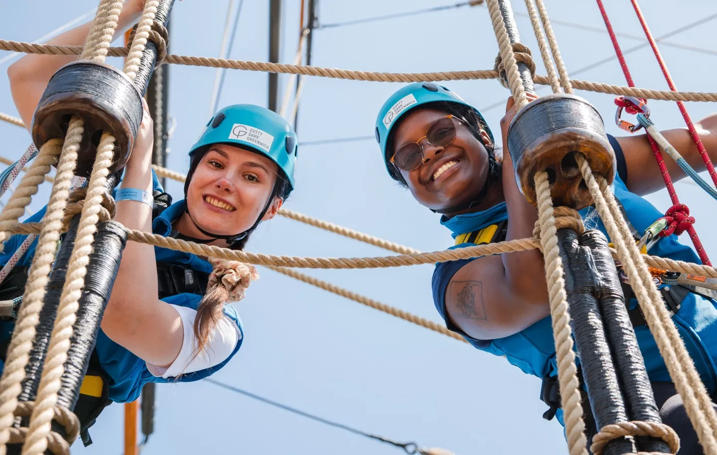 Photo of two women climbing rigging of a historic ship and looking down at the camera whilst smiling
