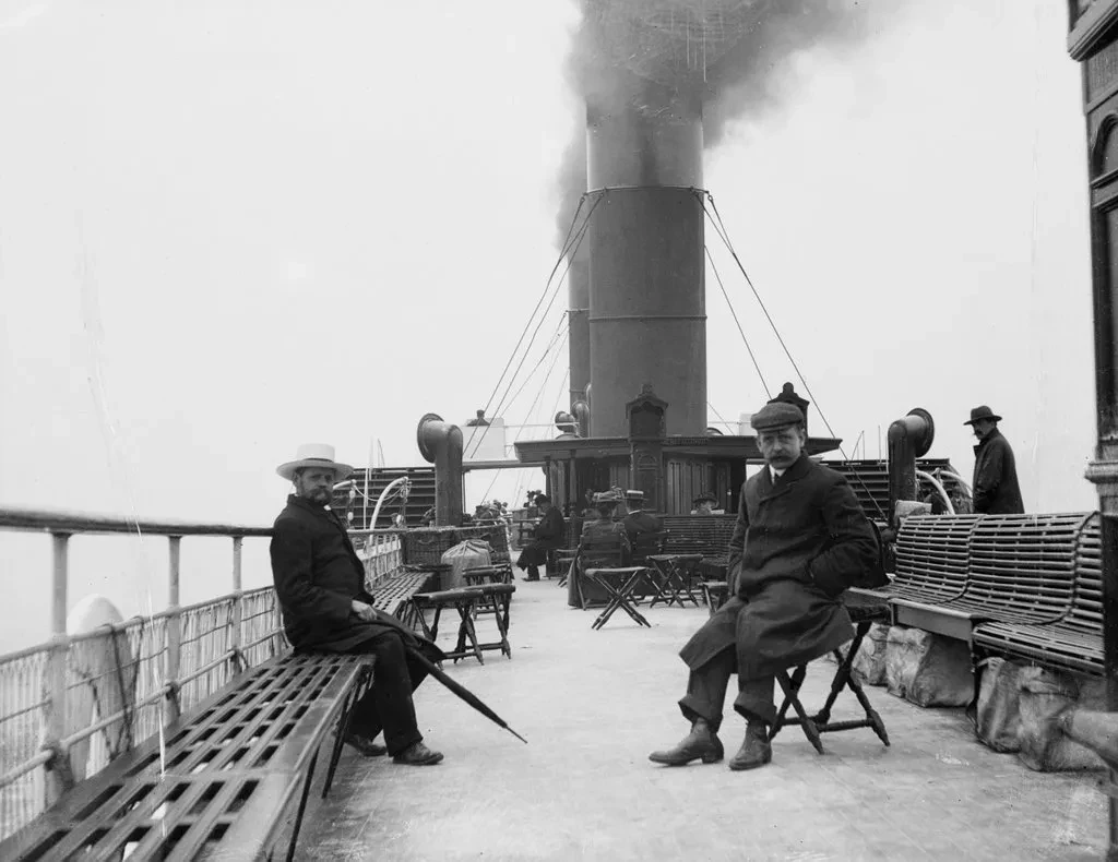 Historic black and white photograph taken on board the Columba paddle steamer. Two men are sat on deck looking towards the camera. Thick black smoke is rising out of the ship's funnels