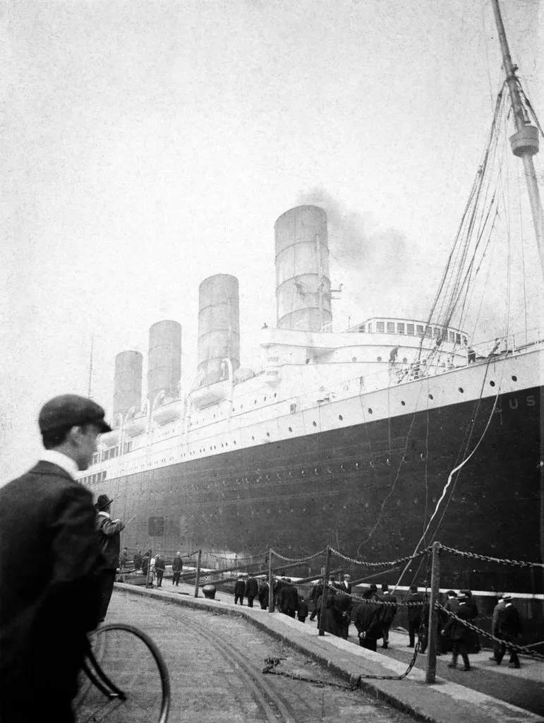 Historic black and white photograph of RMS Lusitania docked in a harbour. A man in a flat cap with his back to the camera is visible in the foreground