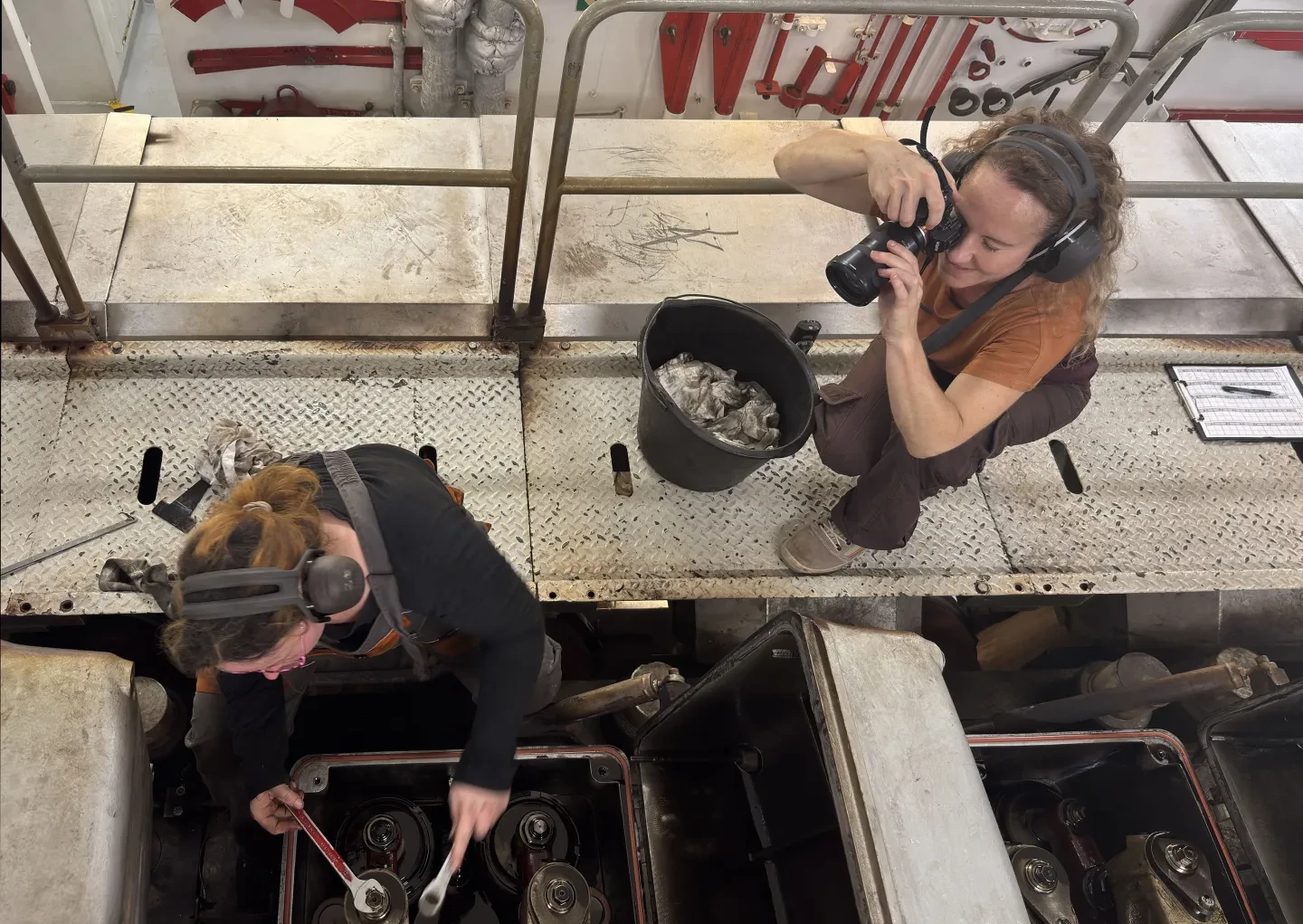 An overhead view showing a photographer kneeling down to take a photo of a female seafarer, who is making repairs to a piece of equipment on deck