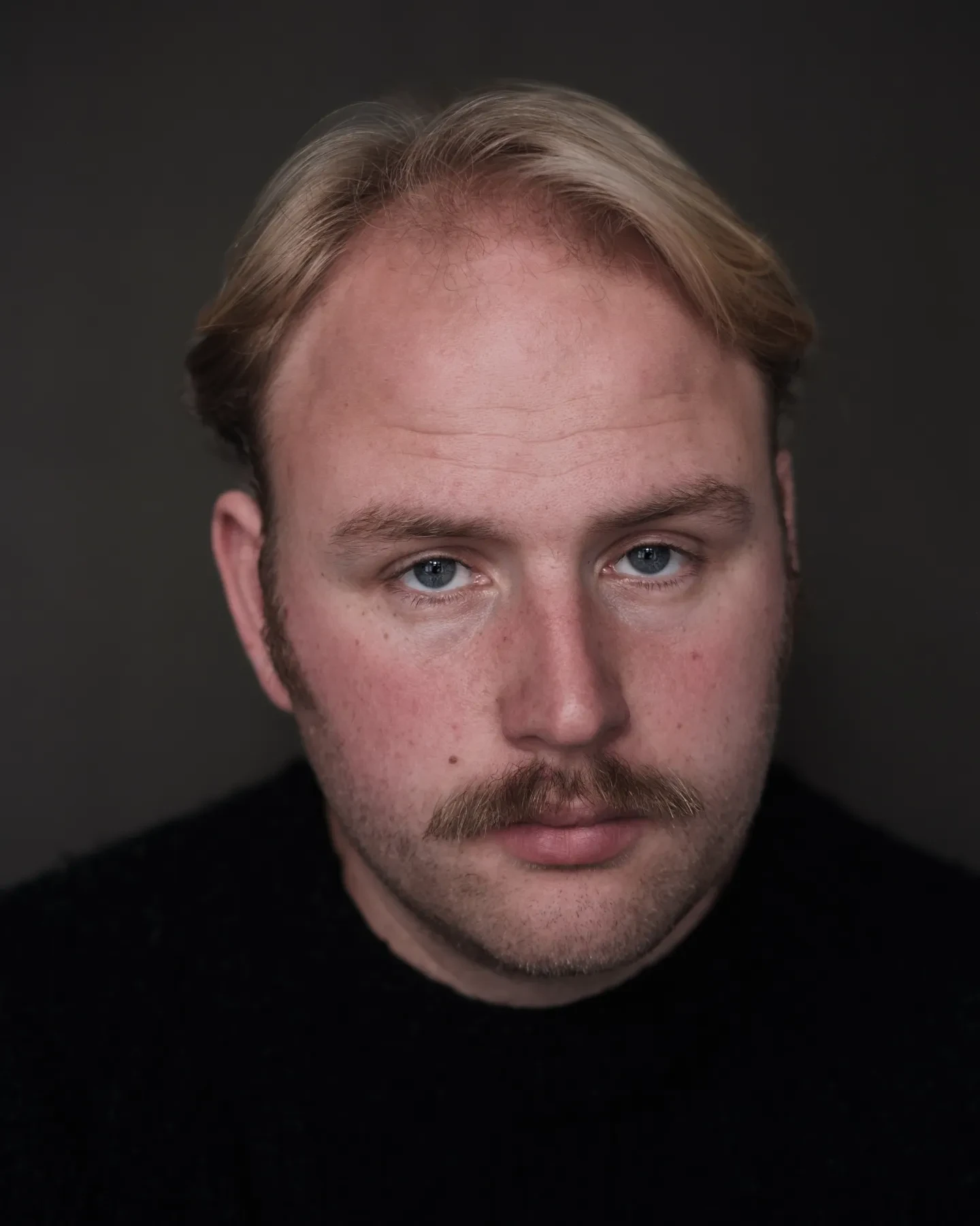 Positioned face on, blonde short hair with moustache, wearing a black high neck top 