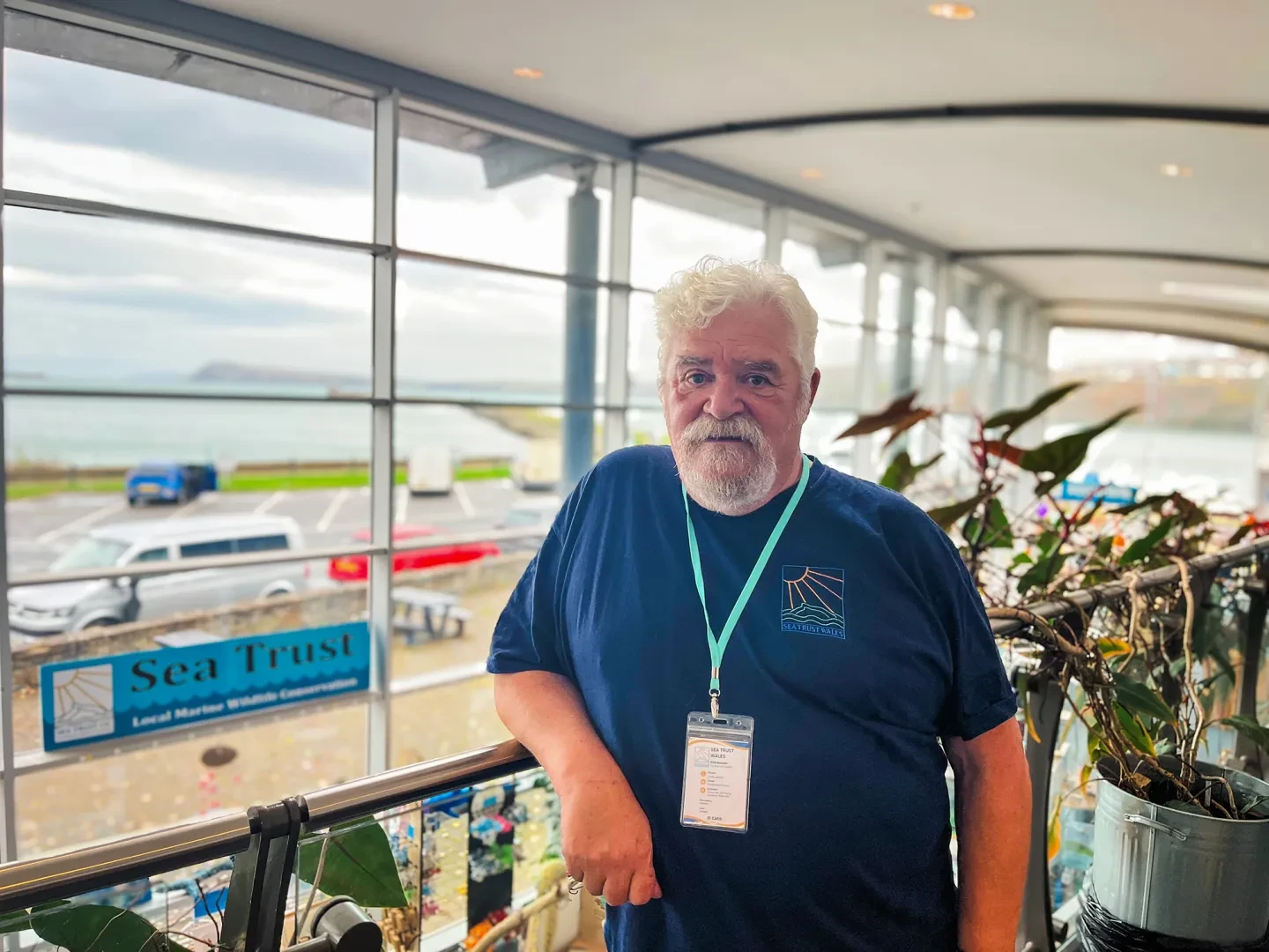 A man stands on an indoor balcony wearing a blue top. Large windows in the background look out on the sea, and the sign 'Sea Trust' is visible on the left hand side
