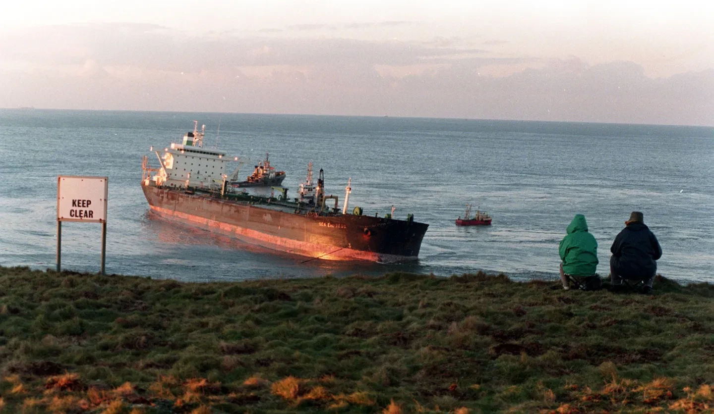 A stricken oil tanker run aground, seen from the coast. Two figures are sitting by the cliff, looking out at the ship, and a sign to their left reads 'KEEP CLEAR'