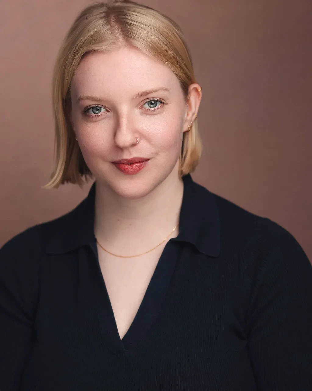 headshot of Verity, position head on towards the camera, smiling, blonde hair, wearing a black v neck shirt with thin gold necklace