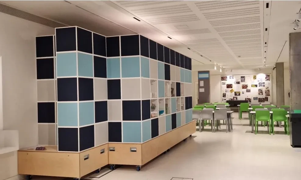 Bright, modern room set up for activities, featuring a large cube installation wall and tables with green chairs for group use.