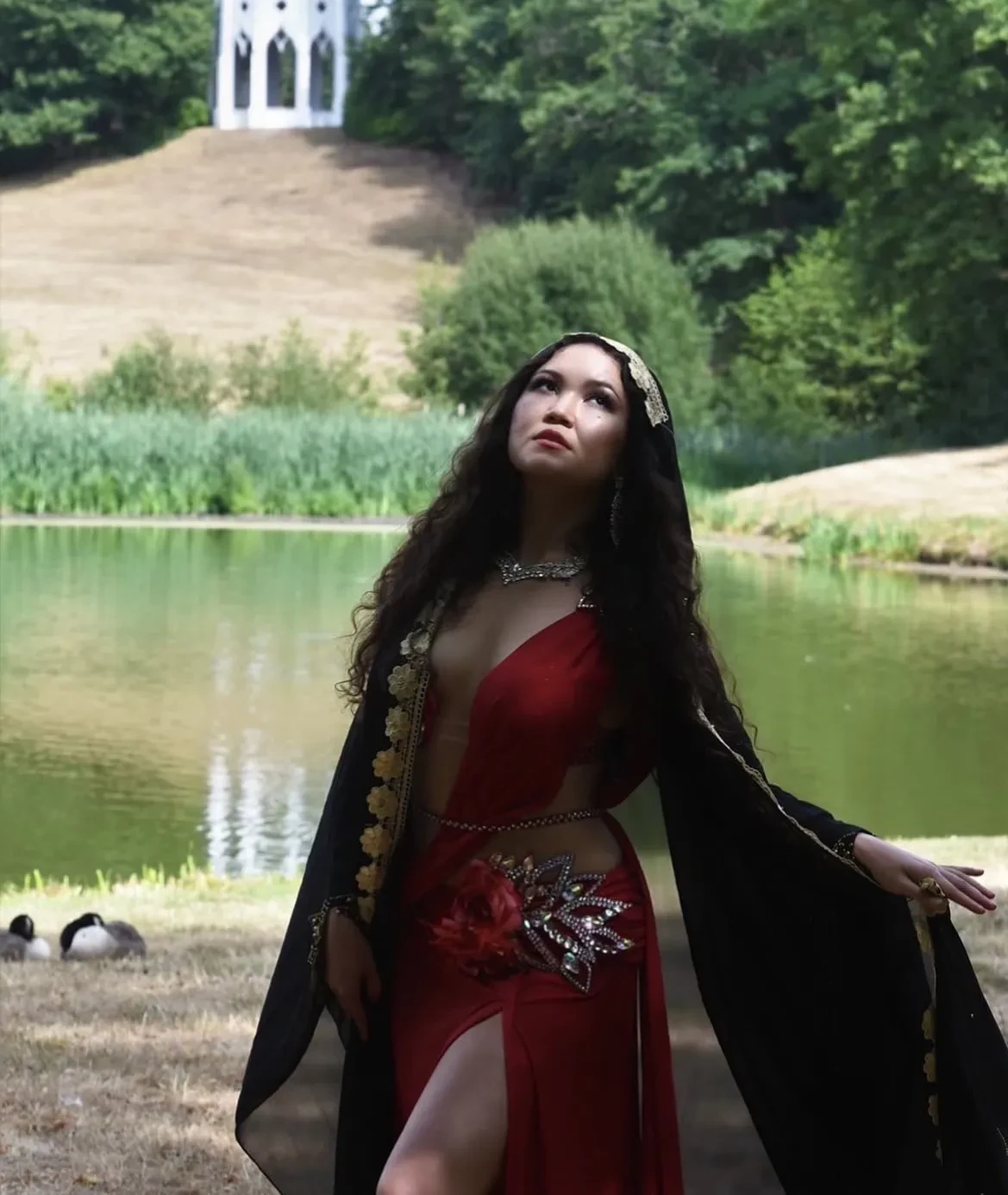 Colour photo with a woman with long, flowing black hair in the forefront and a lake and sloping hill in the background.