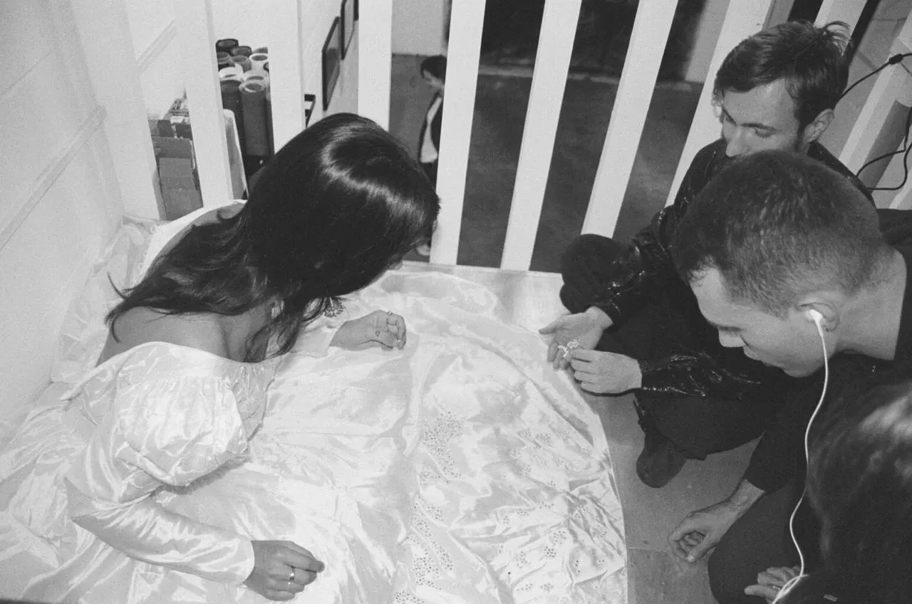 Photo of person wearing white dress which two people are crouching down and embroidering on