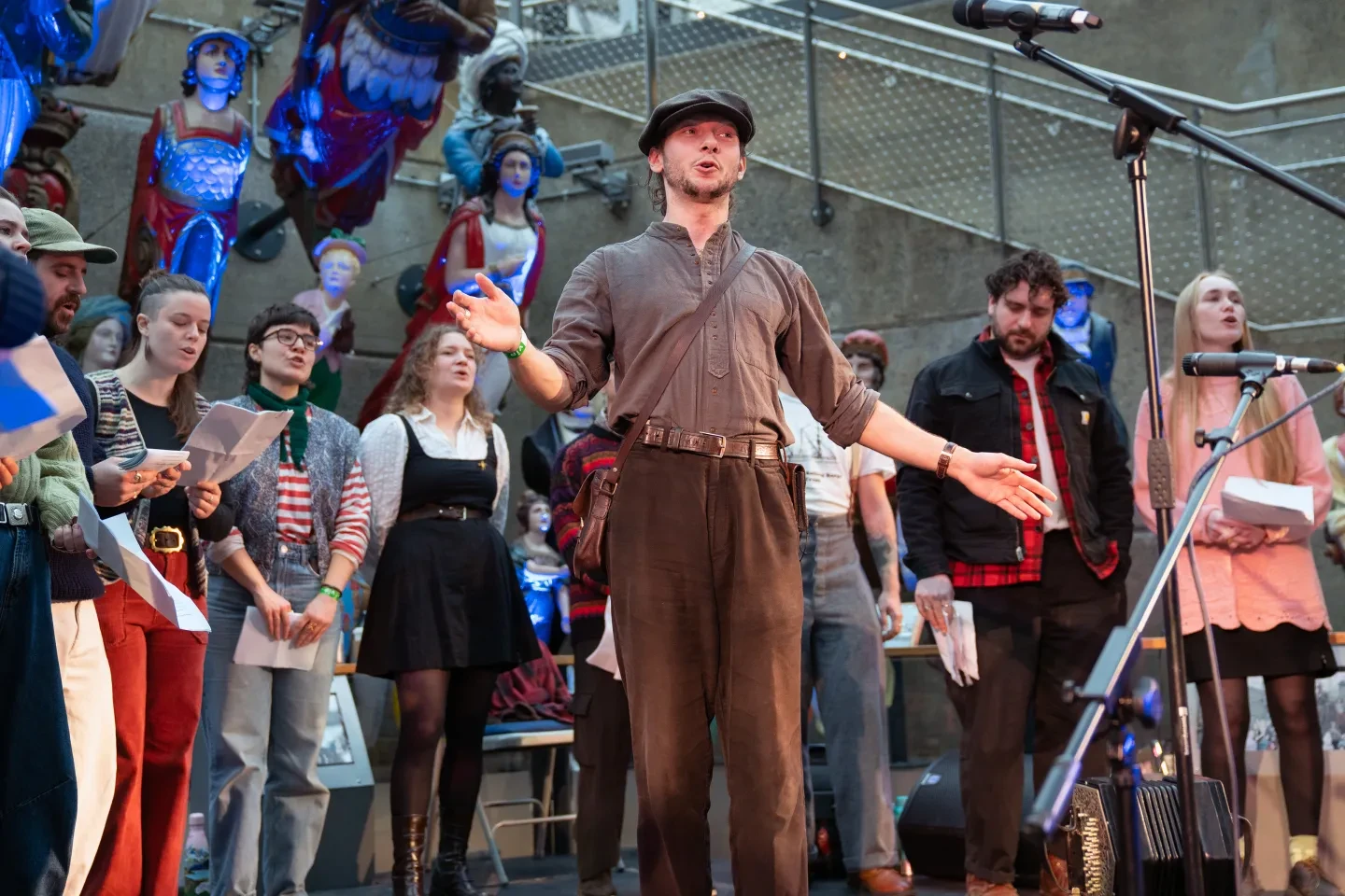 A singer in brown trousers, a brown shirt and a flat cap performs with a choir behind
