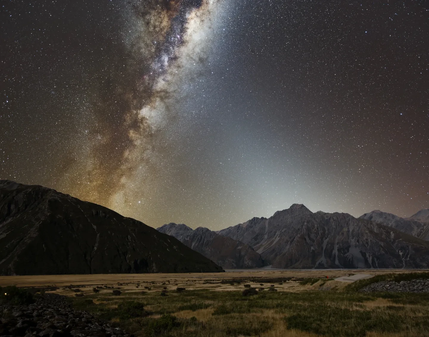 A photo showing a landscape and the night sky above. The night sky includes the Milky Way and a fainter white glow which is zodiacal light.