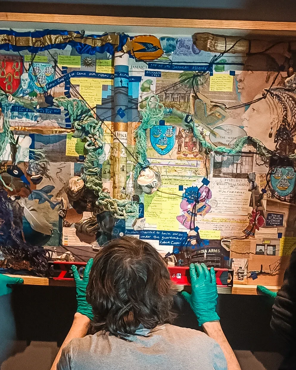 Photo from behind of a man preparing to hang an artwork on a wall. He is kneeling at the bottom of the piece, wearing latex gloves and using a spirit level to check the piece is straight