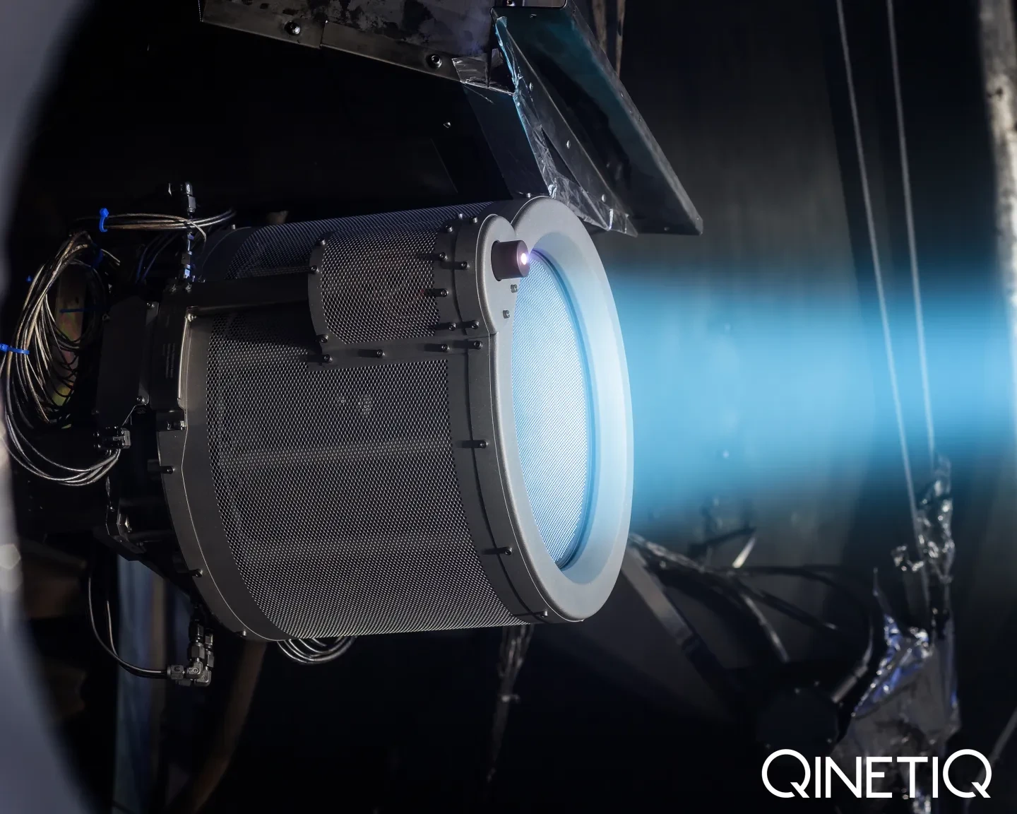 A close up of an ion thruster which appears to be attached to a spacecraft. A metallic drum shape with glowing blue light emanating from it. 