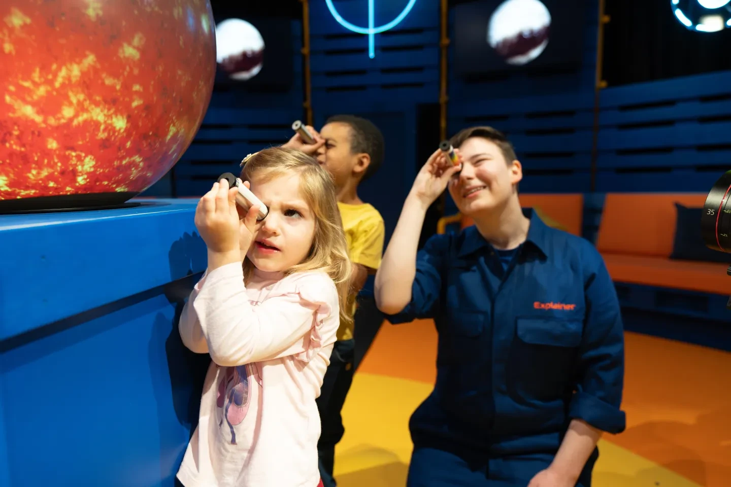 Two young children and an astronomer look through a spectroscope in the Star Huddle, a zone in the Astronomers Take Over experience at the National Maritime Museum