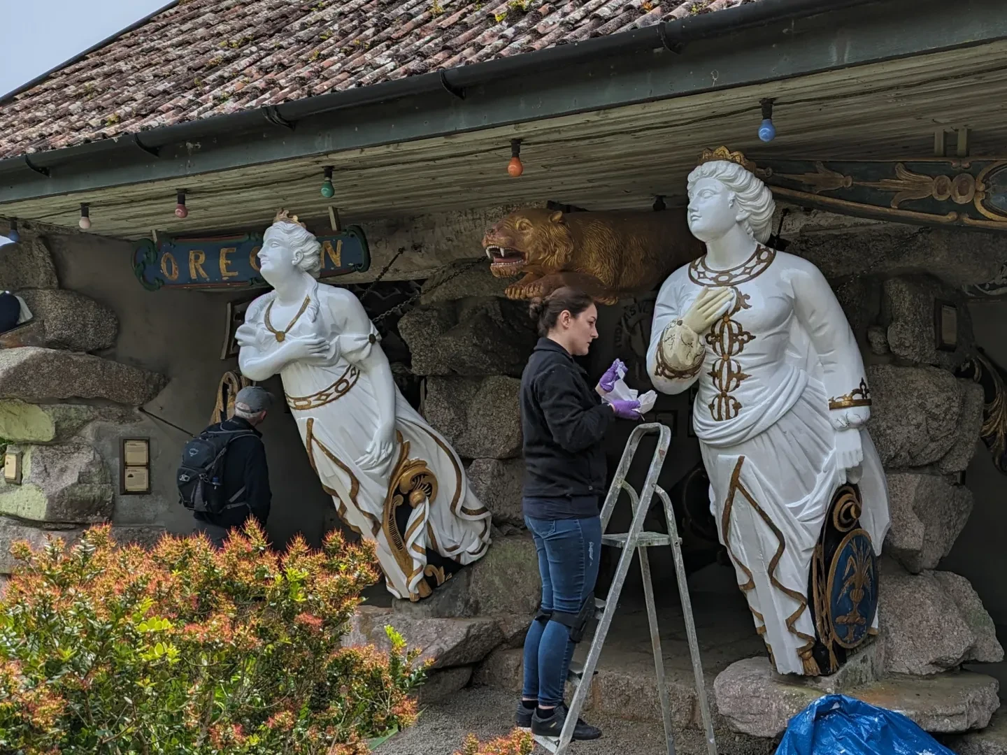 A woman wearing purple gloves stands on a ladder restoring a white figurehead with an intricately carved face and body