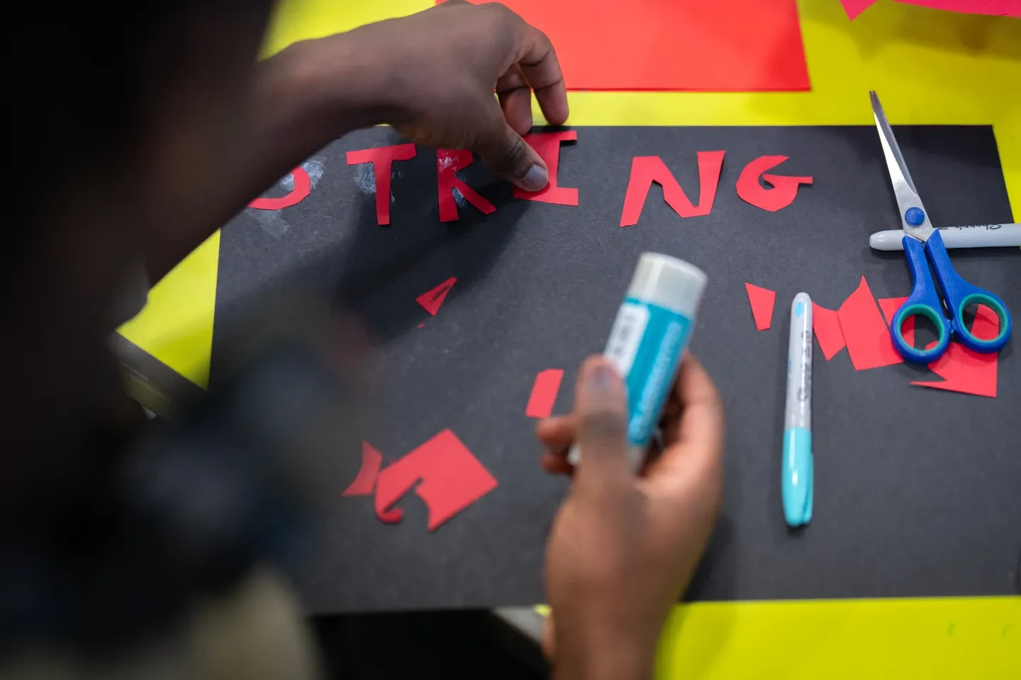 A young person uses glue to stick red letters onto a piece of black card