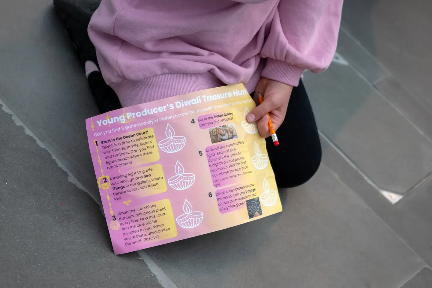 A child in a pink sweatshirt holds a colorful “Young Producer’s Diwali Treasure Hunt” worksheet from the last Young Producers programme and a pencil while sitting on the floor.