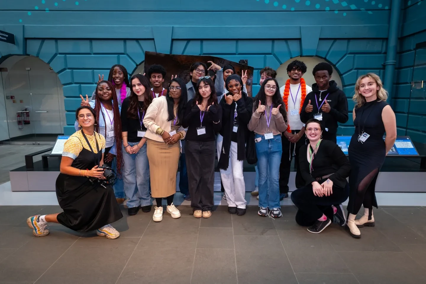 A group of young people and two adults pose together indoors, smiling and gesturing at the camera in a museum-like setting