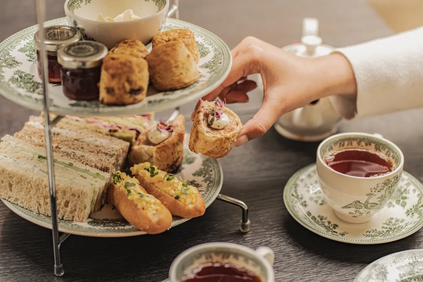 A woman takes a roll from a tiered plate of little sandwiches, with scones, cream and jam on top, and pretty cup of tea on the table