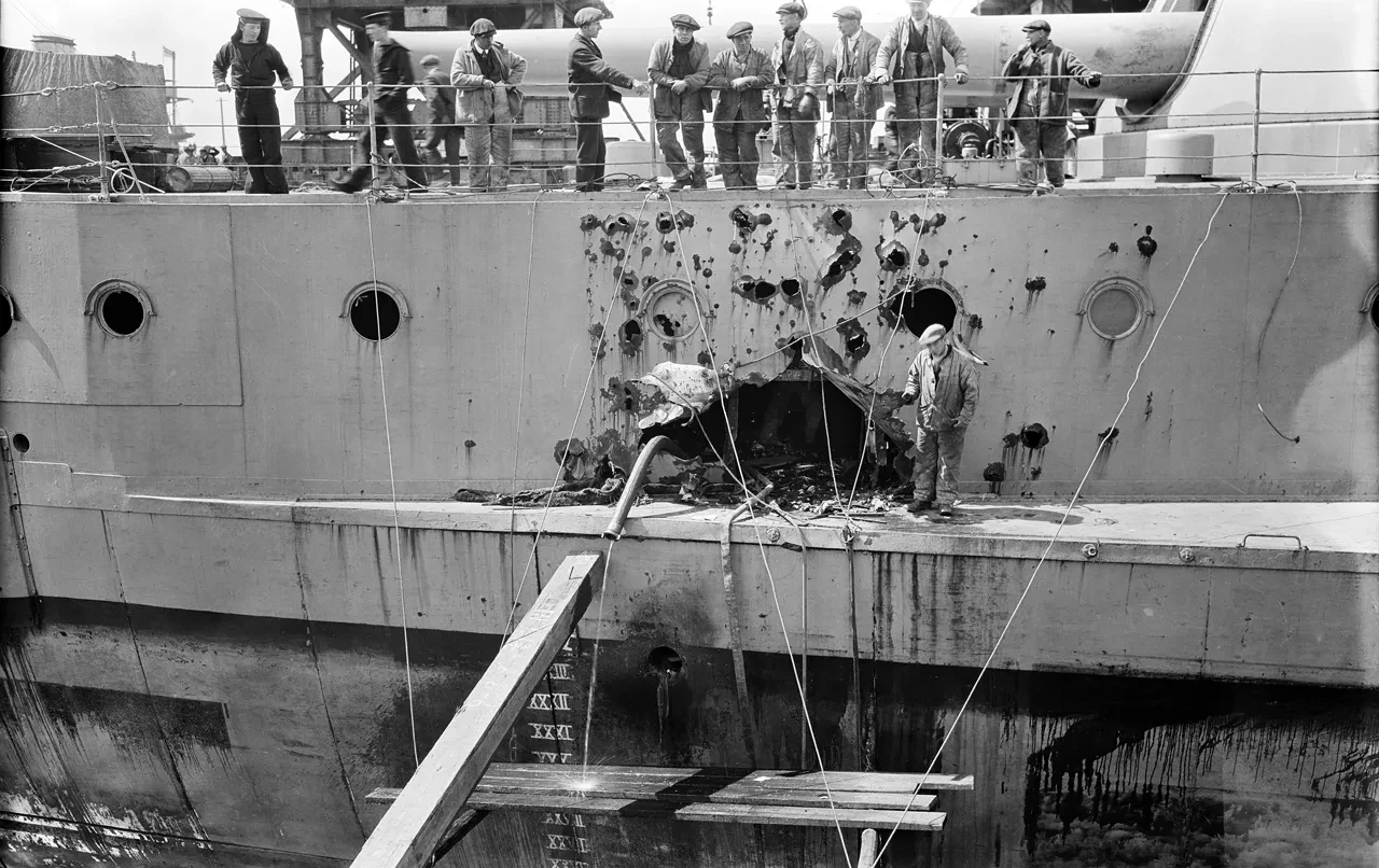 A black-and-white photo of a battleship with shell damage across the side, with a dozen sailors looking at the scarring over the side