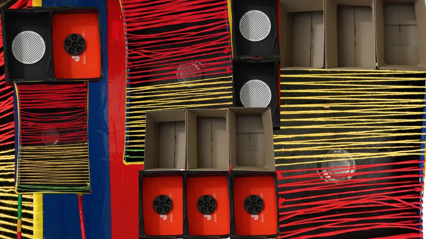 Overhead view of a colorful handmade installation featuring cardboard boxes, black and red panels, circular speaker-like shapes, and tightly stretched red and yellow yarn creating geometric patterns.