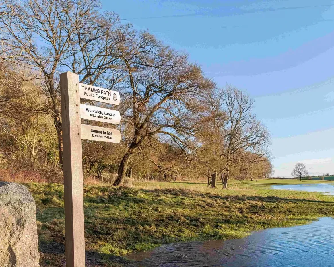 A signpost positioned next to trees and a floodplain. Three arrows point the same direction, reading 'THAMES PATH: Public Footpath', 'Woolwich, London: 185.2 miles 298km', 'Source to Sea: 232 miles 374 km'