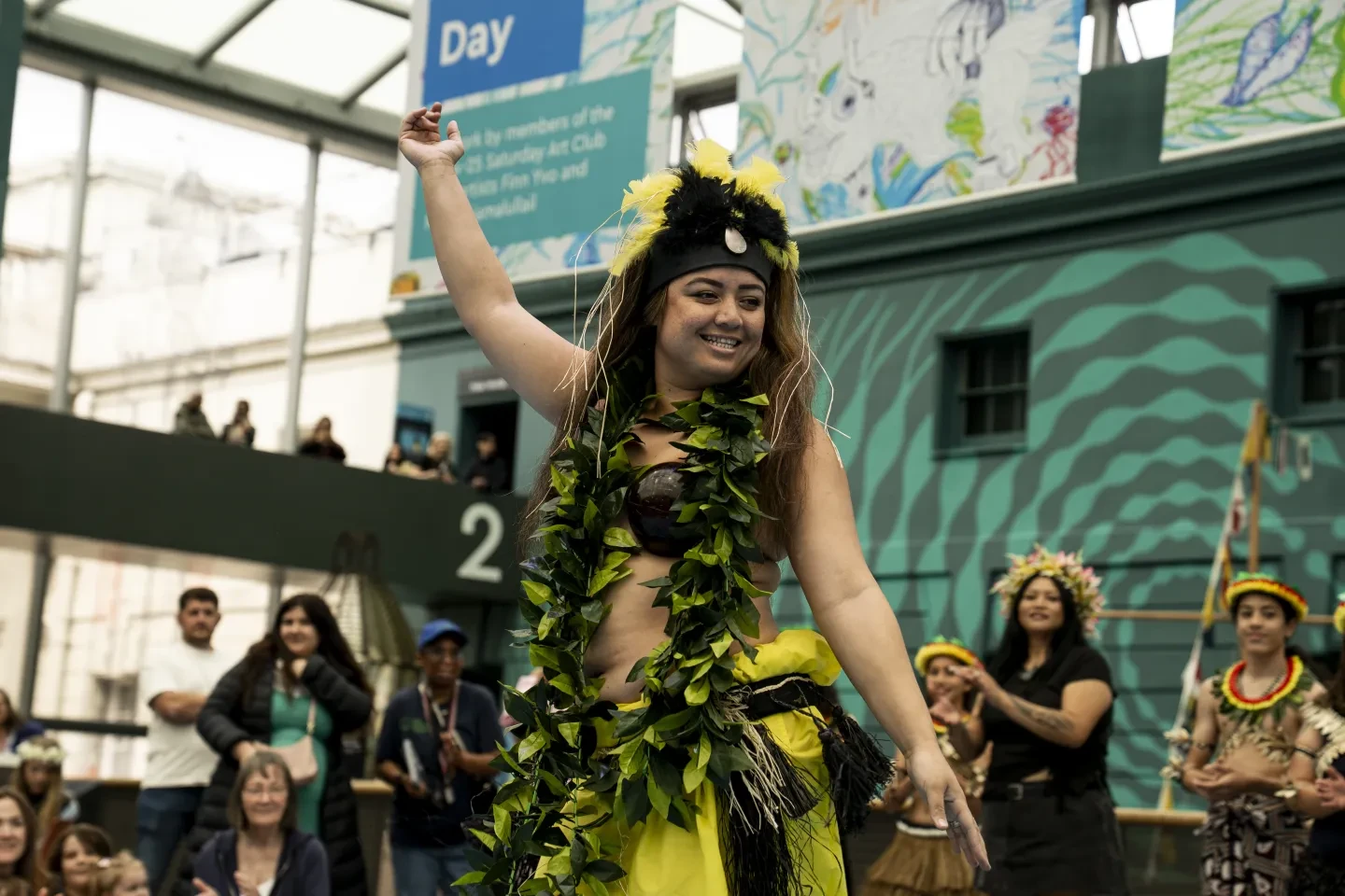 Photo of woman dancing with one arm in the air, wearing a long ley made of leaves, yellow and black headdress and yellow skirt. She is in a large atrium with a crowd of people behind her