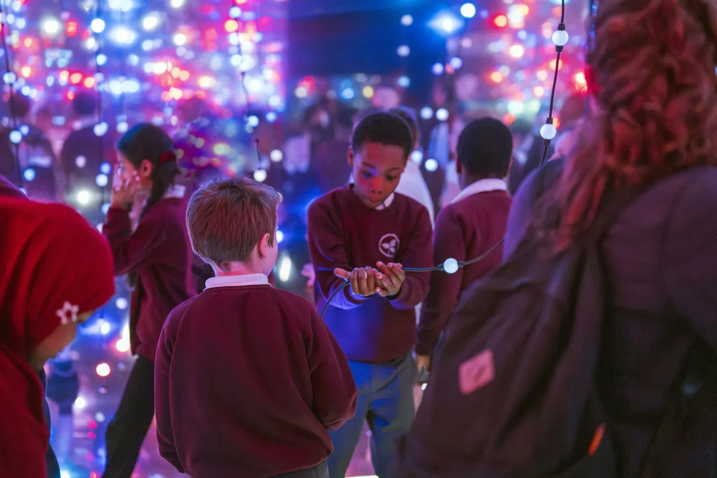 School students standing in a mirrored art exhibition, surrounded by strings of red and blue lights.