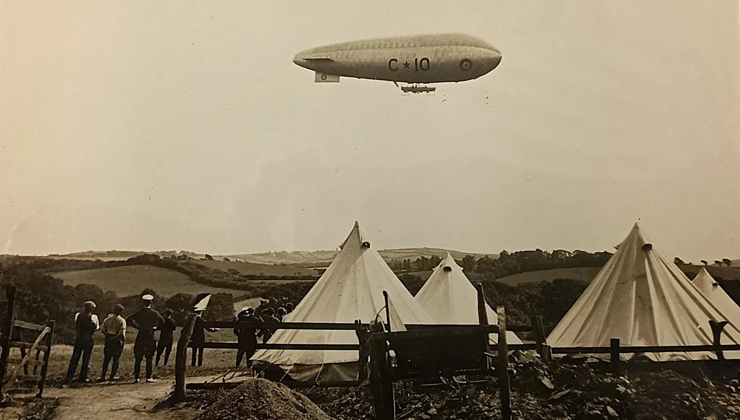 Old photograph of an airship flying over Cornwall. There are some tends and horses in the foreground below..
