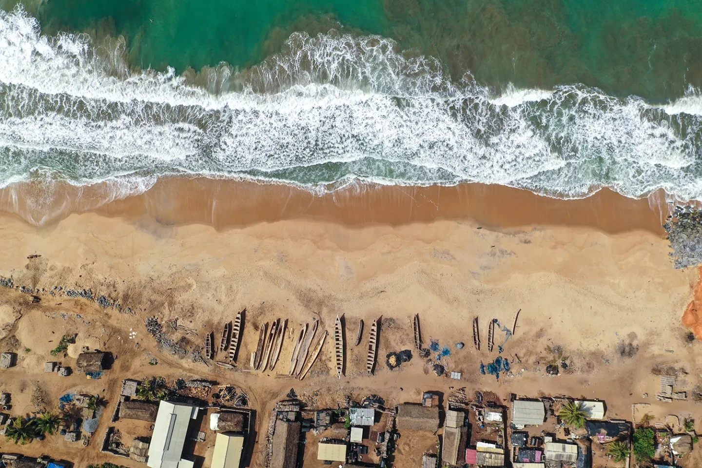 Aerial view of a coastline, featuring waves rolling onto a beach and a line of fishing boats and small huts along the shore
