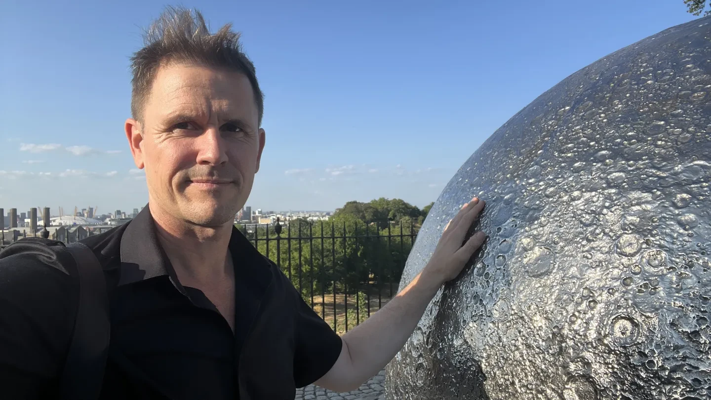 A man in a black shirt stands smiling and touching the surface of a silver Moon sculpture