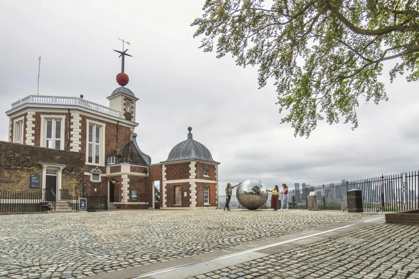 The silver Mirror Moon sculpture in the courtyard at the Royal Observatory in Greenwich, framed by a tree and Flamsteed House, with a grey sky in the background 